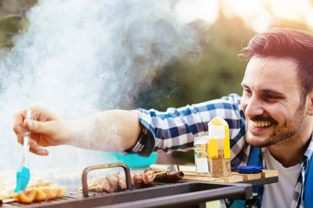 Ένας χαρούμενος άντρας ψήνει κρέας στη σχάρα σε εξωτερικό χώρο, απολαμβάνοντας μια ηλιόλουστη μέρα.