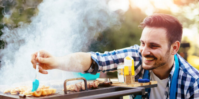 Ένας χαρούμενος άντρας ψήνει κρέας στη σχάρα σε εξωτερικό χώρο, απολαμβάνοντας μια ηλιόλουστη μέρα.