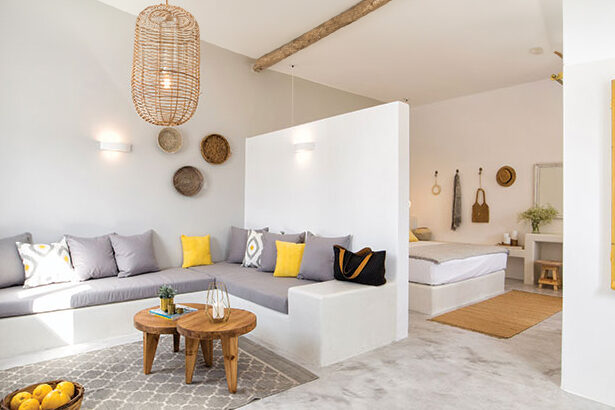 Ένα μοντέρνο και φωτεινό καθιστικό με γκρι και κίτρινα μαξιλάρια, ξύλινο τραπεζάκι και κρεμαστό φωτιστικό, με θέα σε ένα υπνοδωμάτιο στο βάθος.