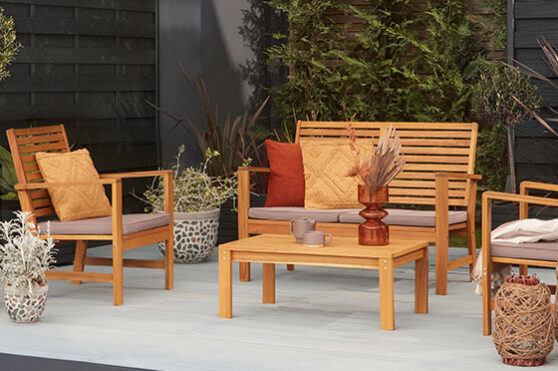 Ένα υπαίθριο καθιστικό με ξύλινα έπιπλα και διακοσμητικά μαξιλάρια, περιτριγυρισμένο από γλάστρες με φυτά.