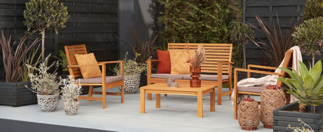 Ένα υπαίθριο καθιστικό με ξύλινα έπιπλα και διακοσμητικά μαξιλάρια, περιτριγυρισμένο από γλάστρες με φυτά.