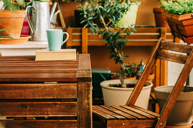 Μικρό ξύλινο τραπέζι και καρέκλα σε μπαλκόνι, περιτριγυρισμένα από γλάστρες με φυτά. Στο τραπέζι υπάρχει μια κούπα και ένα βιβλίο, δημιουργώντας μια ζεστή και χαλαρωτική ατμόσφαιρα.