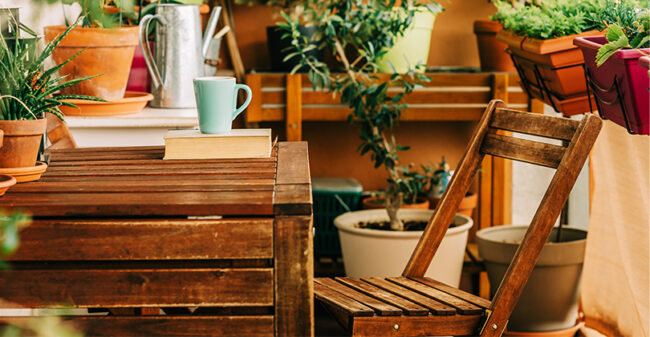 Μικρό ξύλινο τραπέζι και καρέκλα σε μπαλκόνι, περιτριγυρισμένα από γλάστρες με φυτά. Στο τραπέζι υπάρχει μια κούπα και ένα βιβλίο, δημιουργώντας μια ζεστή και χαλαρωτική ατμόσφαιρα.