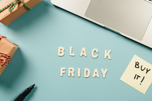 Διαφημιστική εικόνα για τη Black Friday με λέξεις 'Black Friday' γραμμένες σε λευκά γράμματα πάνω σε ανοιχτό γαλάζιο φόντο. Δίπλα υπάρχει ένα αυτοκόλλητο χαρτάκι με τη φράση 'Buy it!' και ένα τυλιγμένο δώρο με κόκκινο και άσπρο κορδόνι. Στην επάνω δεξιά γωνία φαίνεται μέρος ενός φορητού υπολογιστή, ενώ κάτω αριστερά βρίσκεται ένας μαύρος μαρκαδόρος.