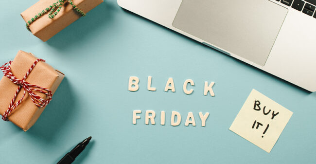 Διαφημιστική εικόνα για τη Black Friday με λέξεις 'Black Friday' γραμμένες σε λευκά γράμματα πάνω σε ανοιχτό γαλάζιο φόντο. Δίπλα υπάρχει ένα αυτοκόλλητο χαρτάκι με τη φράση 'Buy it!' και ένα τυλιγμένο δώρο με κόκκινο και άσπρο κορδόνι. Στην επάνω δεξιά γωνία φαίνεται μέρος ενός φορητού υπολογιστή, ενώ κάτω αριστερά βρίσκεται ένας μαύρος μαρκαδόρος.