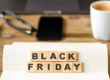 Εικόνα που προωθεί τη Black Friday, με τα γράμματα 'Black Friday' τοποθετημένα σε ξύλινα τετράγωνα πάνω σε ένα τετραδιάκι με καρό σελίδες. Στο υπόβαθρο υπάρχει μια λευκή κούπα καφέ, ένα smartphone, γυαλιά, και ένας φορητός υπολογιστής πάνω σε ξύλινο γραφείο, δημιουργώντας μια αίσθηση οικιακού ή επαγγελματικού περιβάλλοντος.