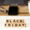 Εικόνα που προωθεί τη Black Friday, με τα γράμματα 'Black Friday' τοποθετημένα σε ξύλινα τετράγωνα πάνω σε ένα τετραδιάκι με καρό σελίδες. Στο υπόβαθρο υπάρχει μια λευκή κούπα καφέ, ένα smartphone, γυαλιά, και ένας φορητός υπολογιστής πάνω σε ξύλινο γραφείο, δημιουργώντας μια αίσθηση οικιακού ή επαγγελματικού περιβάλλοντος.