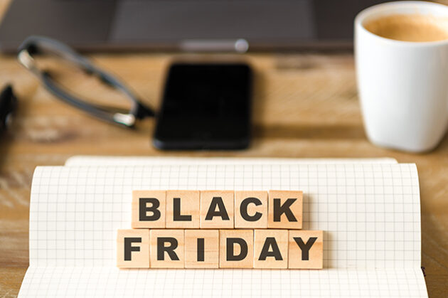 Εικόνα που προωθεί τη Black Friday, με τα γράμματα 'Black Friday' τοποθετημένα σε ξύλινα τετράγωνα πάνω σε ένα τετραδιάκι με καρό σελίδες. Στο υπόβαθρο υπάρχει μια λευκή κούπα καφέ, ένα smartphone, γυαλιά, και ένας φορητός υπολογιστής πάνω σε ξύλινο γραφείο, δημιουργώντας μια αίσθηση οικιακού ή επαγγελματικού περιβάλλοντος.