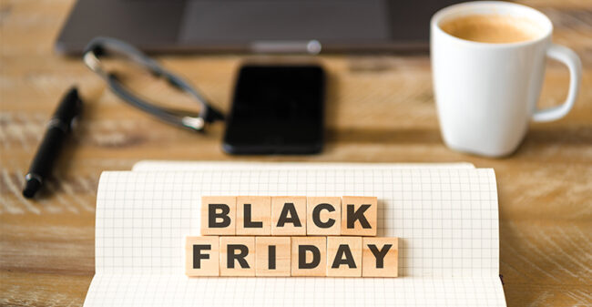 Εικόνα που προωθεί τη Black Friday, με τα γράμματα 'Black Friday' τοποθετημένα σε ξύλινα τετράγωνα πάνω σε ένα τετραδιάκι με καρό σελίδες. Στο υπόβαθρο υπάρχει μια λευκή κούπα καφέ, ένα smartphone, γυαλιά, και ένας φορητός υπολογιστής πάνω σε ξύλινο γραφείο, δημιουργώντας μια αίσθηση οικιακού ή επαγγελματικού περιβάλλοντος.